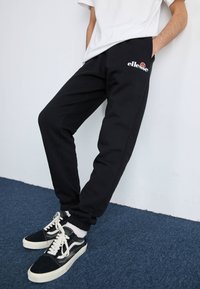 Schwarze Jogginghosen mit elastischem Bund und seitlichen Taschen, mit einem kleinen Logo auf dem linken Oberschenkel und schmal zulaufenden Knöcheln, kombiniert mit schwarzen Sneakern.