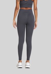 Kobieta w ciemnoszarych legginsach z wysokim stanem i dopasowanym stanikiem sportowym, widoczna z tyłu, stojąca na jednolitym tle.