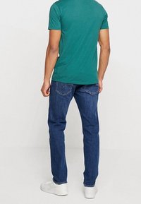 Man wearing teal short-sleeve t-shirt, dark blue jeans, and white sneakers standing facing a plain light background.