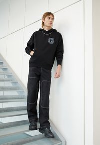 Svart huvtröja med ficka framtill och grafisk logotyp, ihop med svarta byxor med vita sömnadsdetaljer, mot en minimalistisk bakgrund.