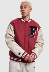 Veste universitaire rouge et crème avec col en tricot, logo "F" brodé, boutons-pression et poignets côtelés rayés. Tissu doux avec des manches contrastantes.