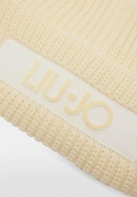 Pletená čiapka v krémovej farbe s textúrovaným povrchom. Obsahuje biely textilný pás s vyrazeným logom "LIU·JO" v zladených krémových odtieňoch.