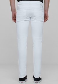 Vaqueros blancos de corte ajustado con una textura suave, que cuentan con dos bolsillos traseros y una cinturilla. Llevados con zapatillas negras.
