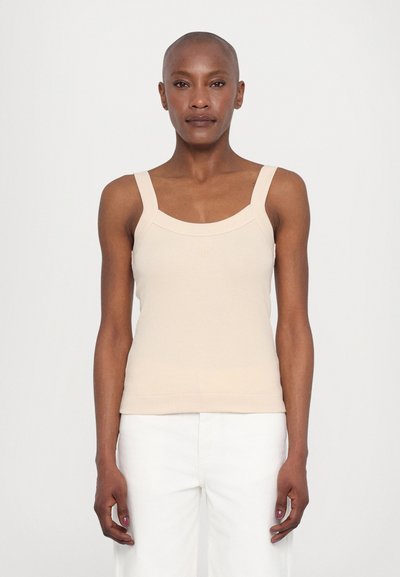 Top de tank beige, cu un model nervurat, cu bretele late și un decolteu în formă de scoică, asortat cu pantaloni albi cu croi larg, având o siluetă ajustată.