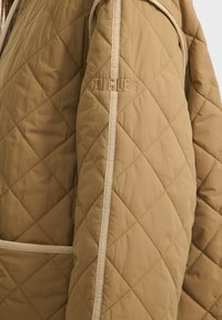 Beige gesteppte Jacke mit diagonalen Nähten, ausgestattet mit einem cremefarbenen Akzentstreifen und gesticktem Logo. Weicher, leichter Stoff.