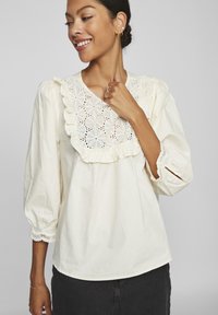 Blouse blanche avec des détails en dentelle festonnée et des découpages floraux au niveau du col. Comprend des manches bouffantes et des bords de bas décoratifs.