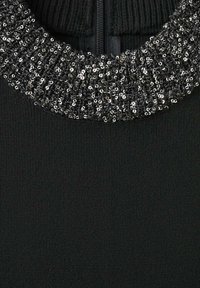 Suéter negro con un cuello texturizado y con cuentas que presenta lentejuelas plateadas. El cuerpo tiene una textura de punto suave y acanalado y un cierre de cremallera.