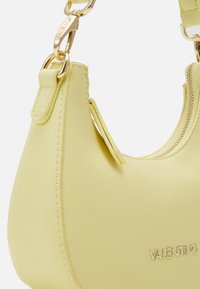Žlutá texturovaná kabelka s zakřiveným tvarem, která má zipové uzavírání a zlaté kovové detaily, s embosovaným názvem "VALENTINO".