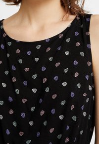 Detailní záběr horní části těla v černém bezrukávovém topu s malými pastelovými vzory listů v růžové, modré, fialové a bílé barvě.