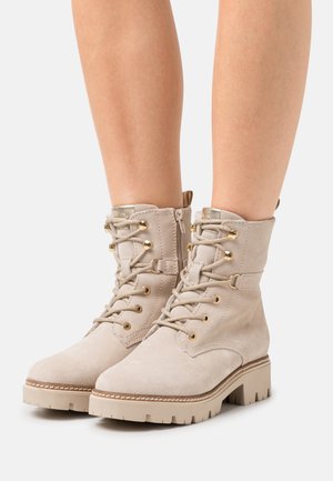 Stivaletti alla caviglia in camoscio beige con lacci, occhielli dorati, cerniera laterale e suole spesse in gomma indossati su gambe nude.