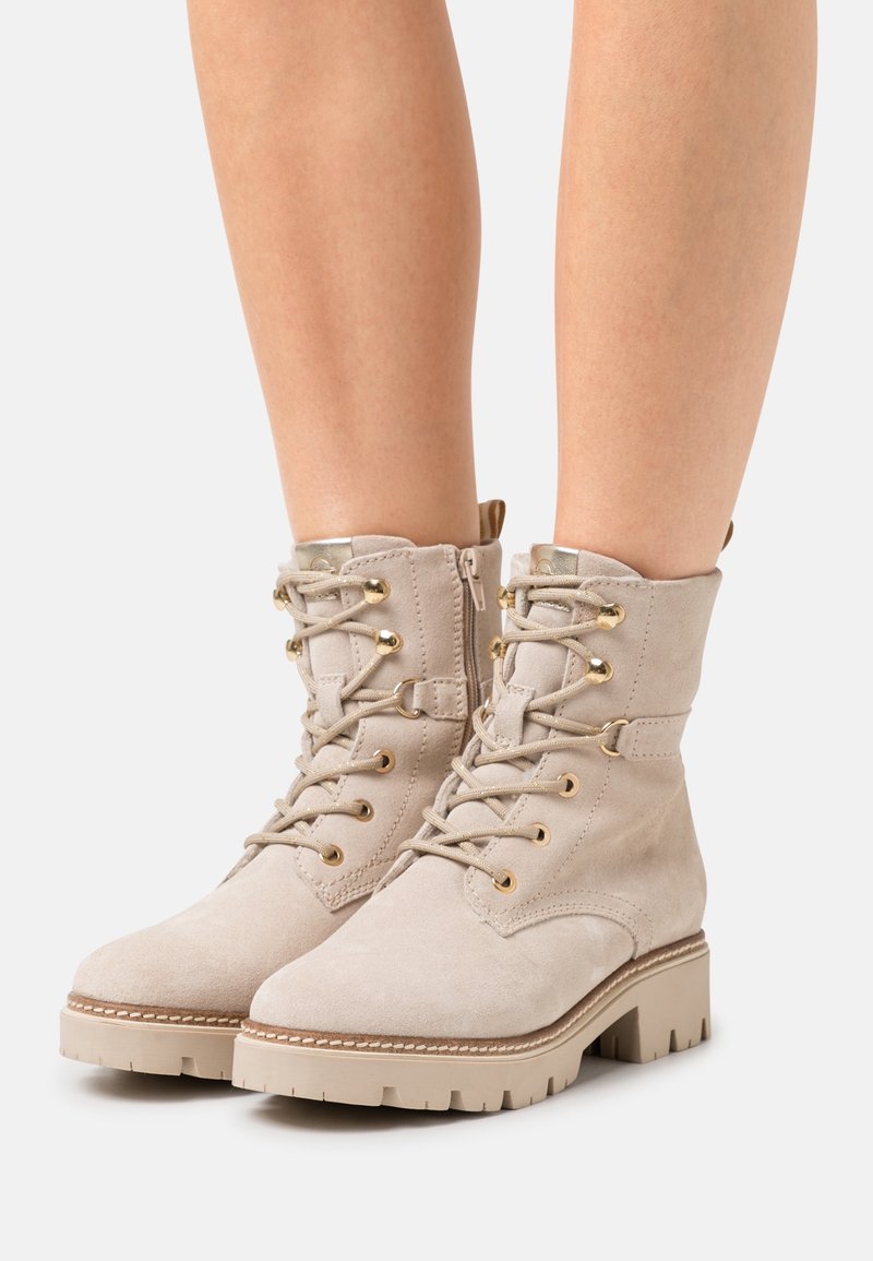 Bottines en daim beige à lacets avec œillets dorés, fermeture éclair latérale et semelles en caoutchouc épaisses portées sur jambes nues.