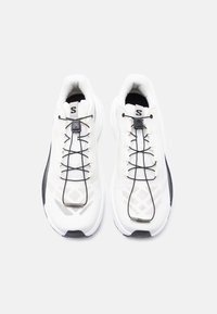 Ζευγάρι λευκών αθλητικών παπουτσιών με μαύρα ελαστικά κορδόνια χωρίς δέσιμο, μαύρες άκρες σόλας και επενδεδυμένα πέλματα, εμφανιζόμενα από πάνω σε λευκό φόντο.