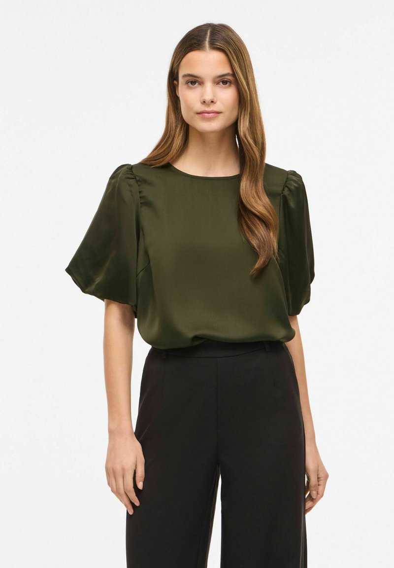 Femme aux longs cheveux bruns portant un chemisier court vert olive à manches bouffantes, rentré dans un pantalon noir taille haute, debout devant un fond uni.