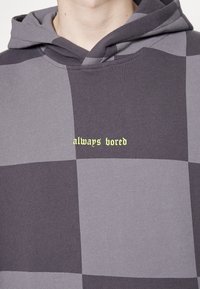 Sudadera con capucha que presenta un patrón de cuadros en tonos de gris con el texto "siempre aburrido" en verde lima en el pecho. Textura suave.