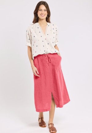 Femme souriante portant un chemisier blanc à manches courtes avec de petits motifs floraux, une jupe midi rose avec une fente devant et des sandales marron.