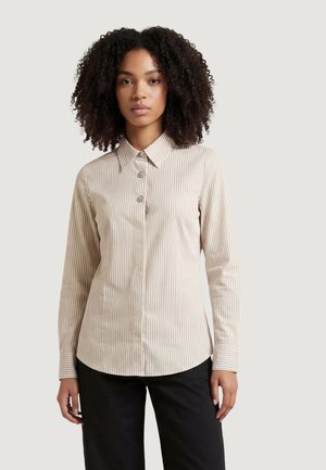 Donna con capelli ricci che indossa una camicia beige a righe bianche con bottoni e pantaloni neri davanti a uno sfondo semplice.