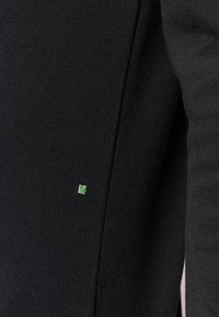 Black fabric garment featuring a textured surface and a small green square accent near the seam. Design is minimalistic and modern.