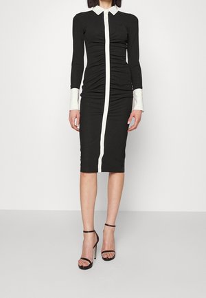 Femme portant une robe midi noire ajustée avec col blanc, poignets blancs et une bande verticale centrale blanche, associée à des sandales à talons noires.