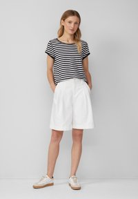 Pasiastrzona czarno-biała bluzka z krótkim rękawem, zestawiona z białymi szortami z plisami oraz beżowymi sneakersami, prezentująca swobodny, luźny styl.
