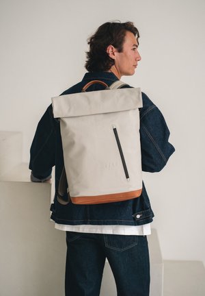 Junger Mann trägt eine dunkelblaue Jeansjacke und Jeans, trägt einen beigen Rolltop-Rucksack mit braunem Boden und seitlichem Reißverschluss und schaut seitlich.