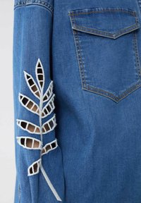 Camicia di jeans in tessuto blu, con un motivo a foglia ricamato in bianco sulla manica, dettagli forati e taschino sul petto.