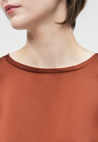 Gintaro spalvos marškiniai su lygiu, matinio paviršiaus tekstūru ir apvaliu iškirpimu. Audinys atrodo lengvas ir laisvai krintantis. Nėra matomų raštų ar aparatūros.