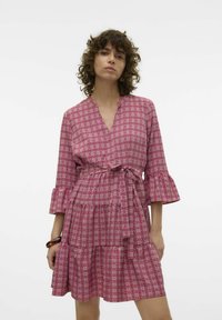 Rozā rakstaina kleita ar V veida kakla izgriezumu, trīs ceturtdaļu zvanu piedurknēm, saitējo vidukli un daudzslāņu ruffles apakšmalu. Izgatavota no viegla auduma.