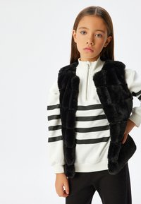 Felpa bianca con righe nere, colletto con zip, abbinata a un gilet senza maniche in pelliccia sintetica nera. La modella indossa pantaloni neri.