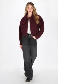 Burgunderfarbene, kürz geschnittene Jacke mit Kragen und Taschen vorne, kombiniert mit locker sitzenden, dunkelgrauen Jeans und schwarzen Schuhen, vor einem weißen Hintergrund.