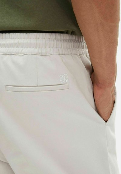 Close-up of a person’s hand in the pocket of light beige pants with elastic waistband and small embroidered logo near back pocket.