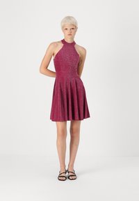 Φόρεμα με λαιμόκοψη halter, μήκους έως το γόνατο, σε μεταλλικό φούξια ύφασμα. Φαρδύ φουστάνι με υφή και εφαρμοστό μπούστο. Μαύρες σανδάλια.