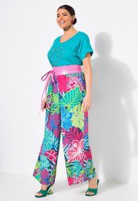 Turquoise T-shirt met korte mouwen en versieringen, gecombineerd met wijde, kleurrijke bloemenpatroonbroekt en een roze riem. Groene hak sandalen.