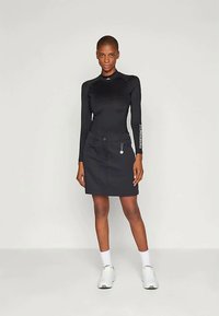 Top negro de manga larga con un diseño ajustado, acompañado de una falda corta negra con un bolsillo frontal y detalles en plateado. Zapatillas blancas completan el conjunto.