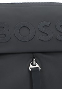 Sac en synthétique noir présentant un logo « BOSS » en relief, fermeture zippée avec tirette métallique et surface texturée. Design minimaliste.