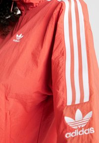 Korálová bunda adidas s zipovým límcem, na rukávech bílými třemi pruhy a logem na hrudi. Lehká, hladká tkanina.