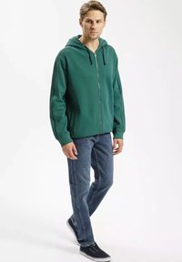 Zielona bluza z kapturem na zamek, z podszewką kaptura, długimi rękawami i kontrastowymi ciemnymi paskami po bokach, w zestawie z niebieskimi dżinsami i czarnymi trampkami.