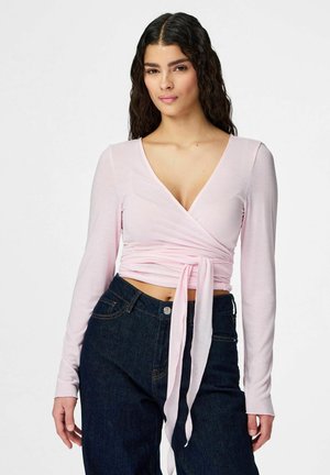 Kvinde med langt krøllet hår iført en lyserød langærmet crop top med omslagsstil og mørke, højtaljede jeans, stående foran en ensfarvet baggrund.