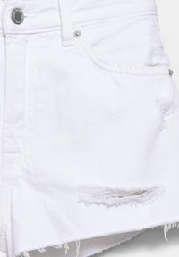 Jupe en denim blanc avec ourlet effiloché et détail déchiré au-dessus du bord inférieur sur le côté avant droit, bouton argenté et rivets visibles.
