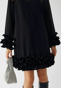 Črna obleka z dolgimi, prosojnimi rokavi in obsežnimi dekorativnimi nagubi na manšetah in robu. Ima preprost V-izrez.