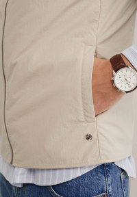 Bežs viegls veste ar gludu tekstūru, ar rāvējslēdzēju, sānu kabatām un logotipa akcentu apakšmalā, pārī ar svītrotu krekls un pulksteni.