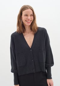 Mörkgrå kort cardigan med djup V-ringning, långa ärmar, två framfickor och fyra stora knappar nedanför mitten. Slät textur.
