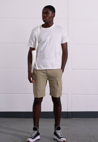 T-shirt en coton blanc, shorts cargo beige avec poches latérales, baskets montantes noir et blanc, et un tote bag blanc. Debout sur un sol en béton.