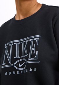 Svart sweatshirt med en framträdande vit och grå "NIKE"-logotyp med "SPORTSWEAR" nedanför, rund halsringning och mjuk textur.