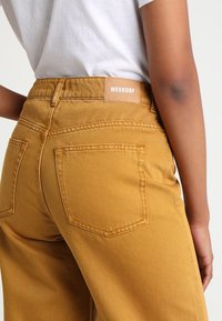 Jeans de algodón amarillos de pierna ancha con cintura alta, que cuentan con bolsillos traseros y una cinturilla etiquetada. Textura suave y costura limpia.