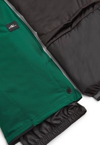 Tejido de chaqueta verde y negra con una textura suave. Presenta puños elásticos, un parche con el logo y bloques de color contrastantes.