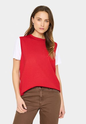 Rode mouwloze top met een ronde hals, gelaagd over een witte shirt met korte mouwen, gecombineerd met bruine getailleerde broek. Eenvoudig en gestructureerd ontwerp.