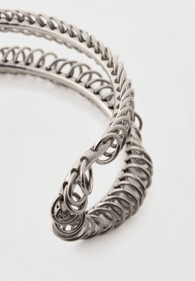 Δύο συνυφασμένα βραχιόλια από ασημένιο μέταλλο με περίπλοκα σχέδια συνδεδεμένων κρίκων σε λευκό φόντο.