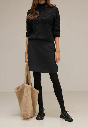 Pull en tricot noir avec un motif en chevrons texturé, associé à une jupe noire. Le modèle tient un sac fourre-tout en polaire beige. Des collants noirs et des chaussures complètent le look.