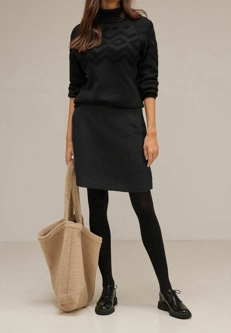 Svart stickad tröja med en strukturerad chevronmönster, i kombination med en svart kjol. Modellen håller en beige fleecetote. Svarta strumpbyxor och skor fulländar looken.
