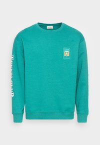 Sweatshirt turquoise à manches longues. Présente un logo graphique sur la poitrine et le texte « TENNIS CLUB » le long de la manche gauche. Texture douce.